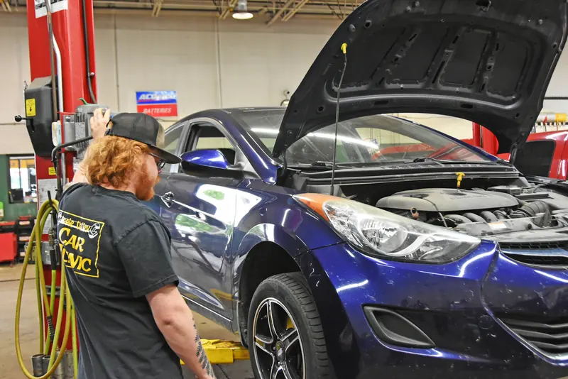 Technician beside a blue sedan with the hood open in the Apple Valley Car Clinic service bay
