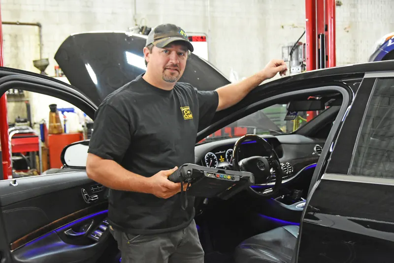 Technician holding a diagnostic scan tablet beside a black luxury sedan